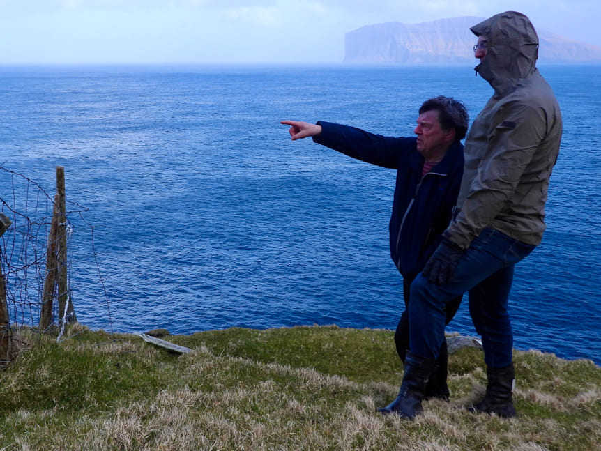 Two men looking at the horizon with the sea in the background, pointing with their fingers.